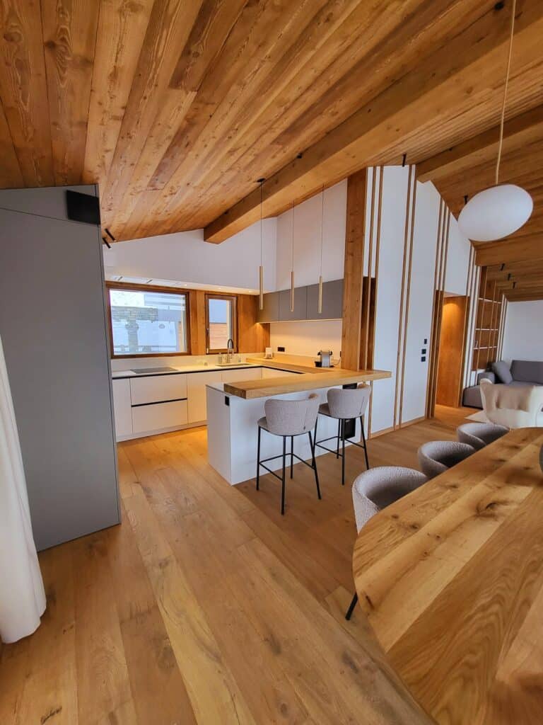 Cuisine moderne ouverte sur salle à manger d'un chalet. Plafond en bois, îlot avec comptoir bois, tabourets et table en bois. Ambiance chaleureuse.