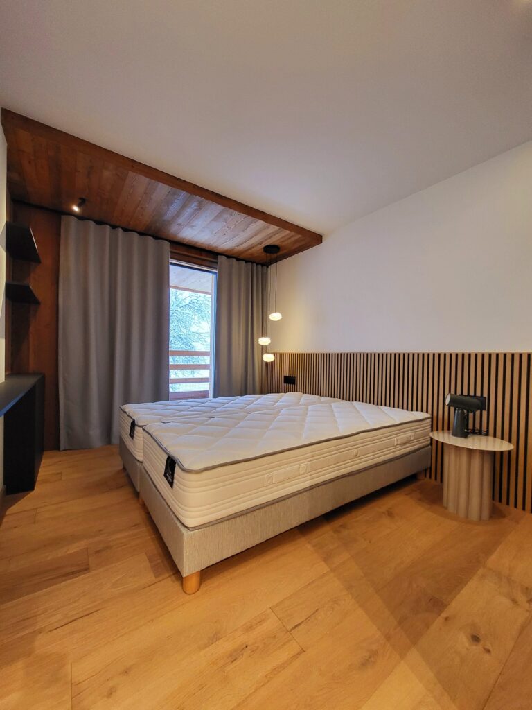 Chambre moderne épurée: lit double confortable, tête de lit en lattes bois clair, parquet et plafond bois asymétrique. Lumière naturelle et suspension design.