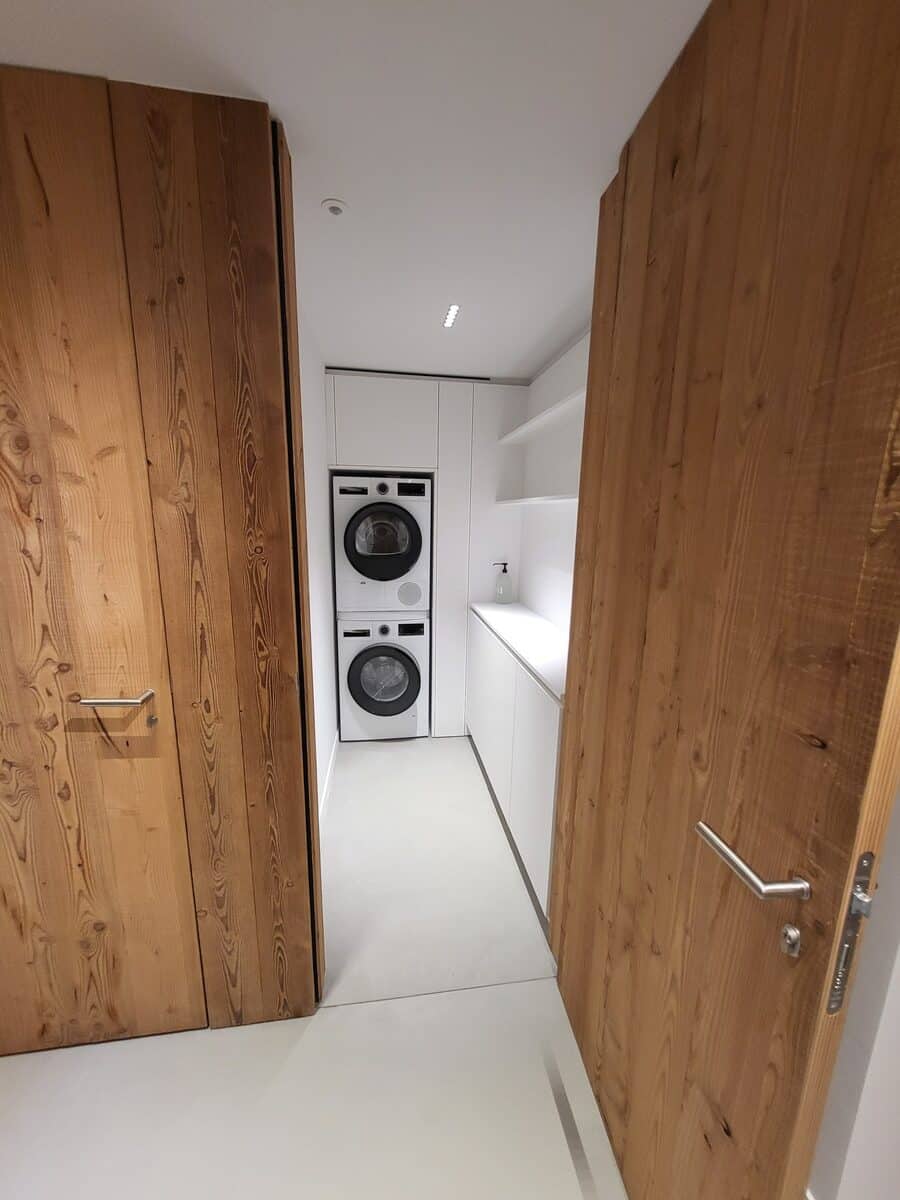 Buanderie moderne et étroite avec lave-linge et sèche-linge superposés, comptoir blanc, étagères, et portes en bois clair ouvertes.