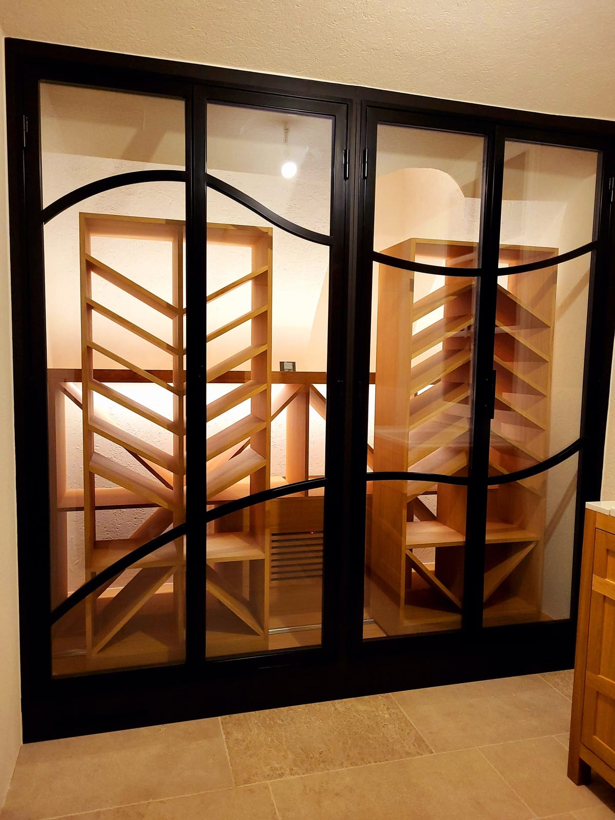 Cave à vin vitrée moderne avec cadre noir, casiers en bois clair et barres métalliques courbes. Éclairée, sol carrelé.