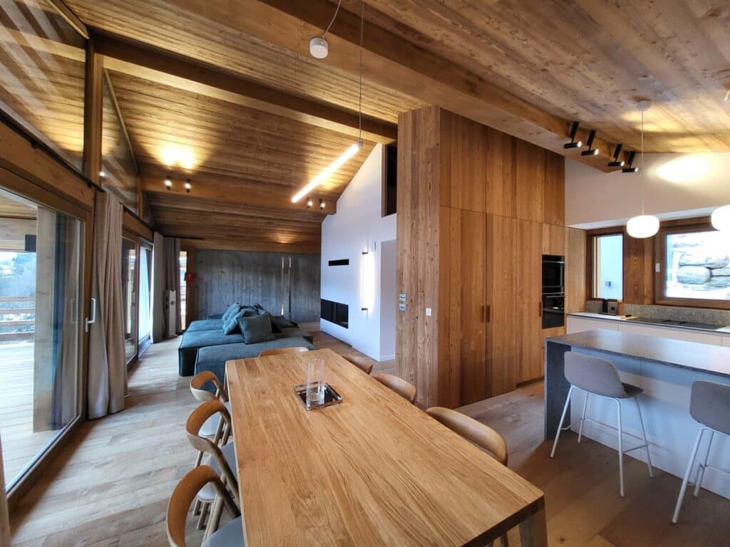 Chalet contemporain en bois: salon avec grand canapé vert, salle à manger et cuisine ouverte, grandes baies vitrées sur un paysage enneigé.