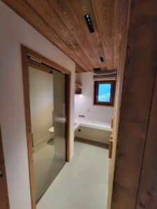 Couloir moderne de chalet. Plafond en bois clair, sol gris. Porte coulissante vitrée vers un WC. Bureau intégré sous une fenêtre donnant sur la neige.