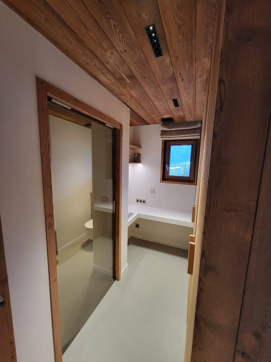 Couloir moderne de chalet. Plafond en bois clair, sol gris. Porte coulissante vitrée vers un WC. Bureau intégré sous une fenêtre donnant sur la neige.
