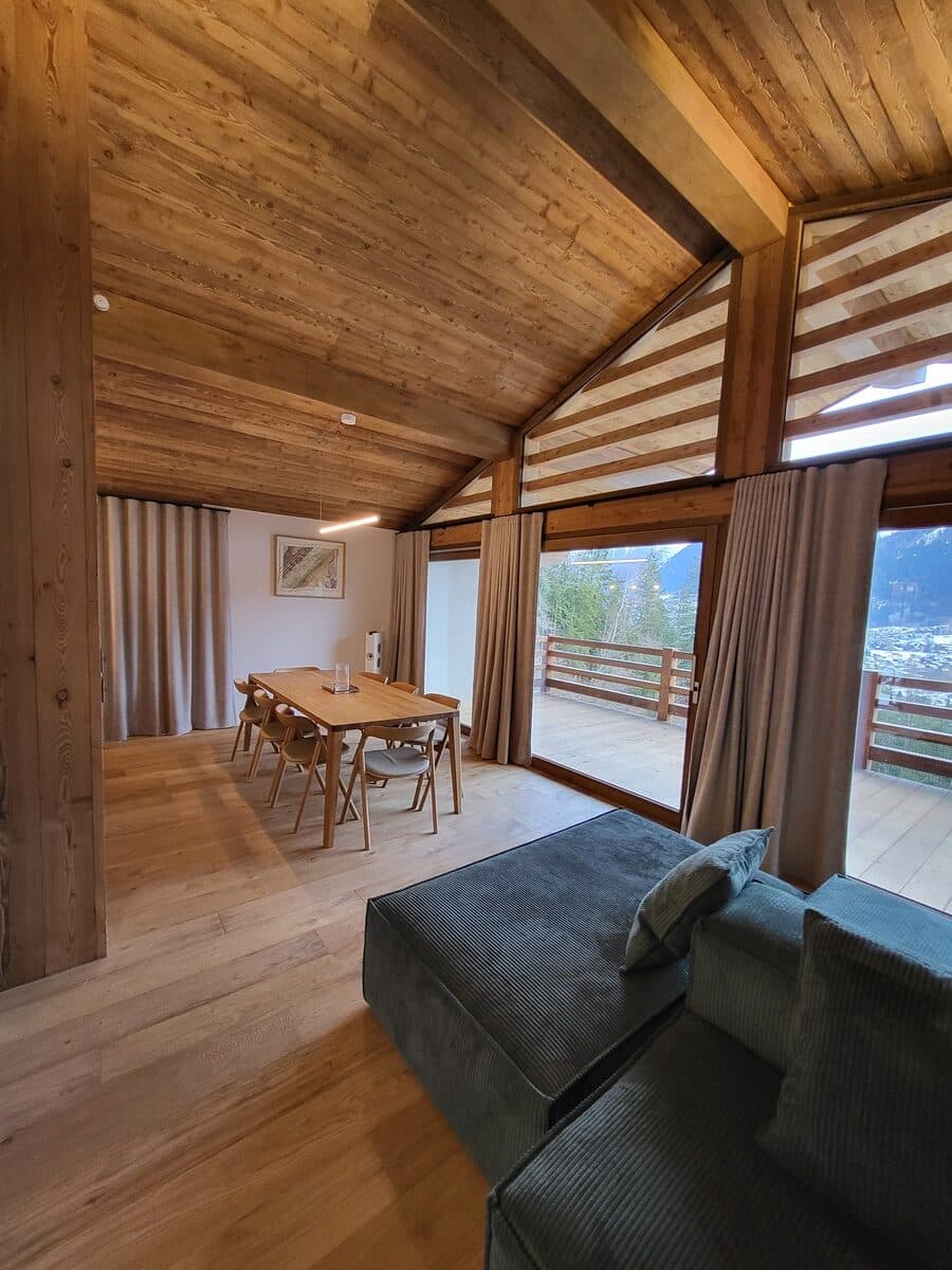 Intérieur de chalet moderne avec plafond et sol en bois clair. Salle à manger en bois, canapé côtelé bleu-gris, vue montagne, terrasse.