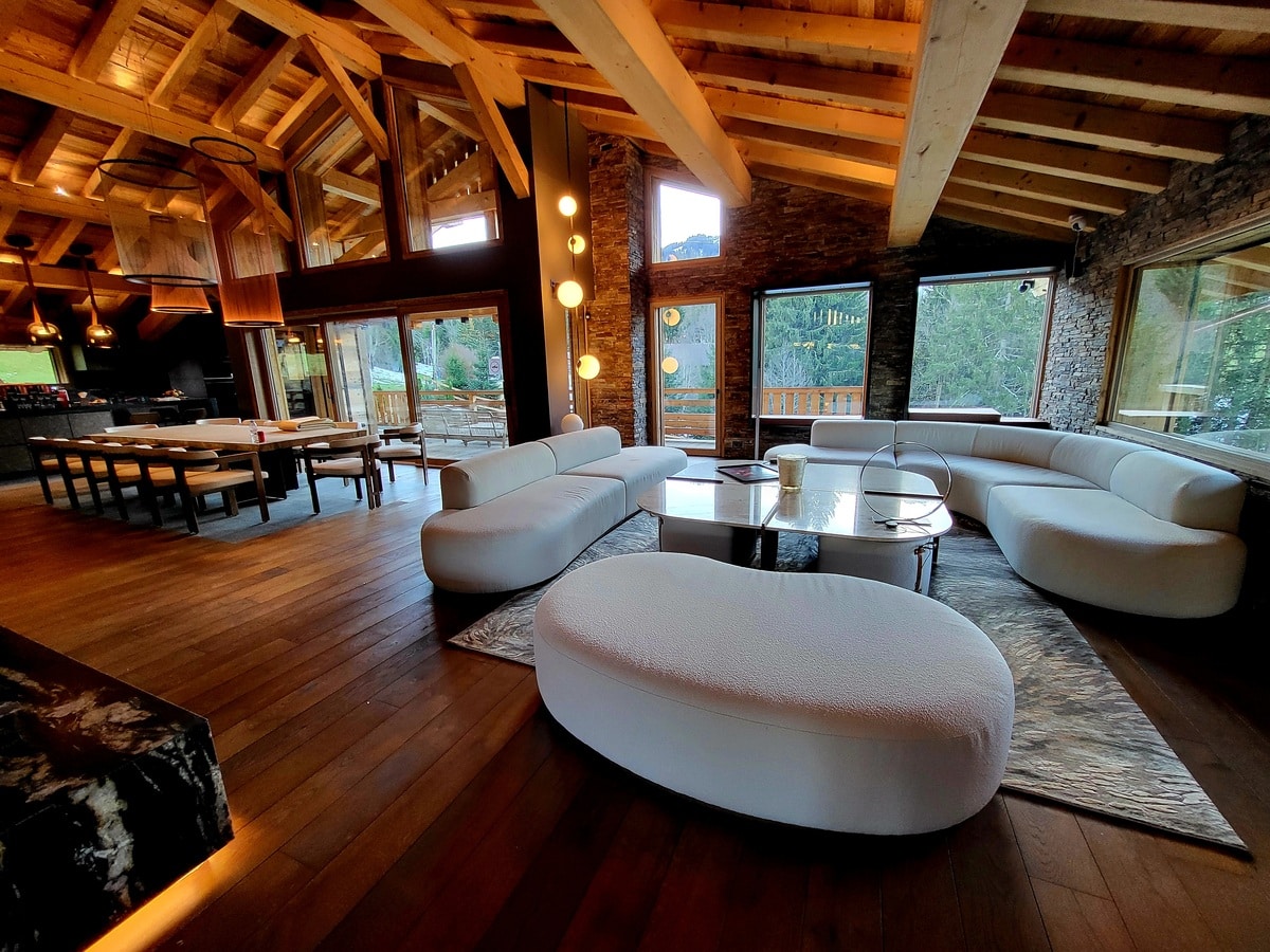 Luxueux chalet moderne : salon design avec canapé blanc incurvé, table basse, et salle à manger en bois. Vue sur forêt enneigée.