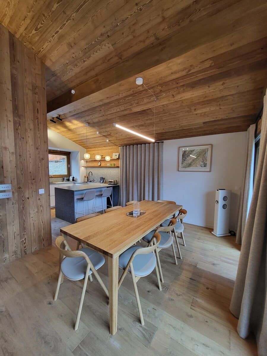 Salle à manger et cuisine ouverte design. Grande table en chêne, chaises grises, plafond et murs en bois clair. Lumière linéaire moderne, ambiance chalet contemporaine.