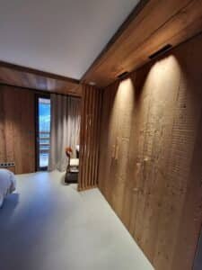 Chambre design en bois avec murs lambrissés, éclairage spots intégré et grand dressing. Sol gris moderne et porte-fenêtre vers la nature.