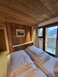 Chambre d'hôtel style chalet avec murs et plafond en bois. Deux lits simples blancs, un tableau de montagne, et un balcon vue montagne enneigée.