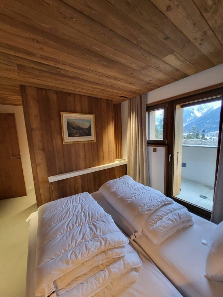 Chambre d'hôtel style chalet avec murs et plafond en bois. Deux lits simples blancs, un tableau de montagne, et un balcon vue montagne enneigée.