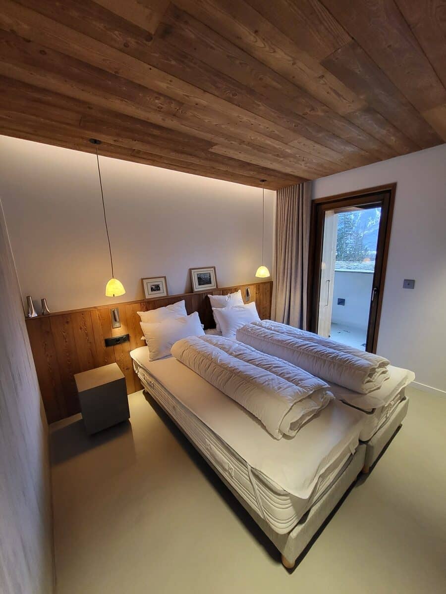 Chambre d'hôtel alpine moderne. Plafond et tête de lit en bois naturel, deux lits confortables, luminaires suspendus. Vue sur un paysage enneigé.