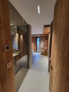 Couloir moderne menant à une chambre d'hôtel en bois clair, avec salle de bain vitrée, sol design et vue montagne.