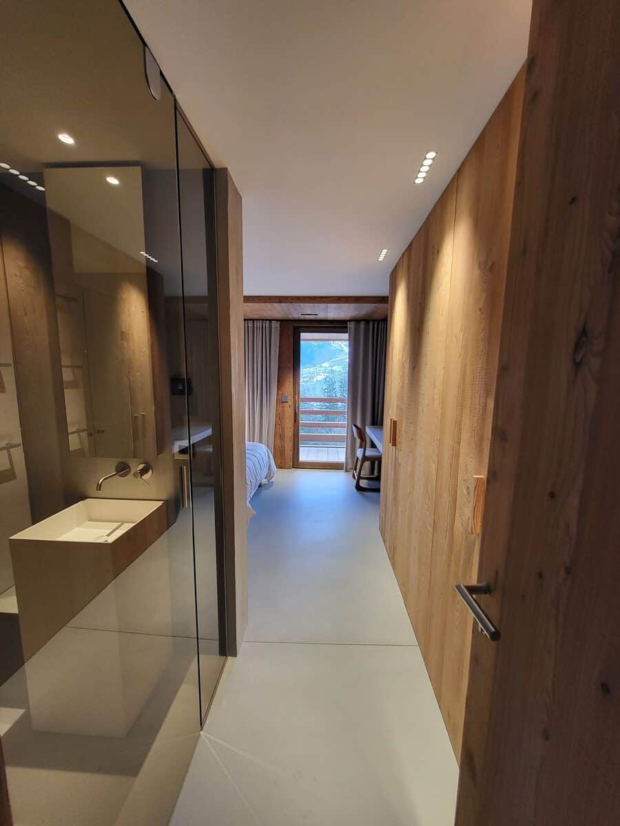 Chambre d'hôtel moderne: salle de bain ouverte, vasque design, vue montagne enneigée. Couloir lambris bois, lumières encastrées.