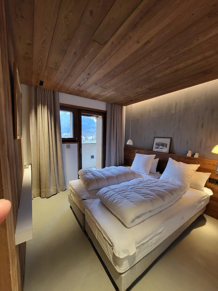 Chambre d'hôtel moderne avec lit double, plafond en bois, vue sur montagnes enneigées. Ambiance chaleureuse et lumineuse.
