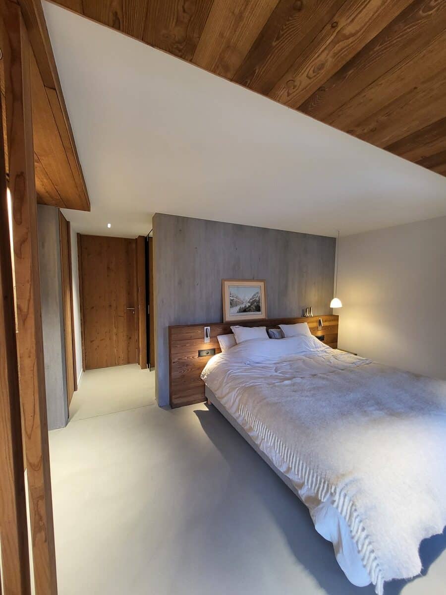 Chambre moderne et chaleureuse avec lit double, tête de lit en bois, mur texturé en béton et plafond bois.
