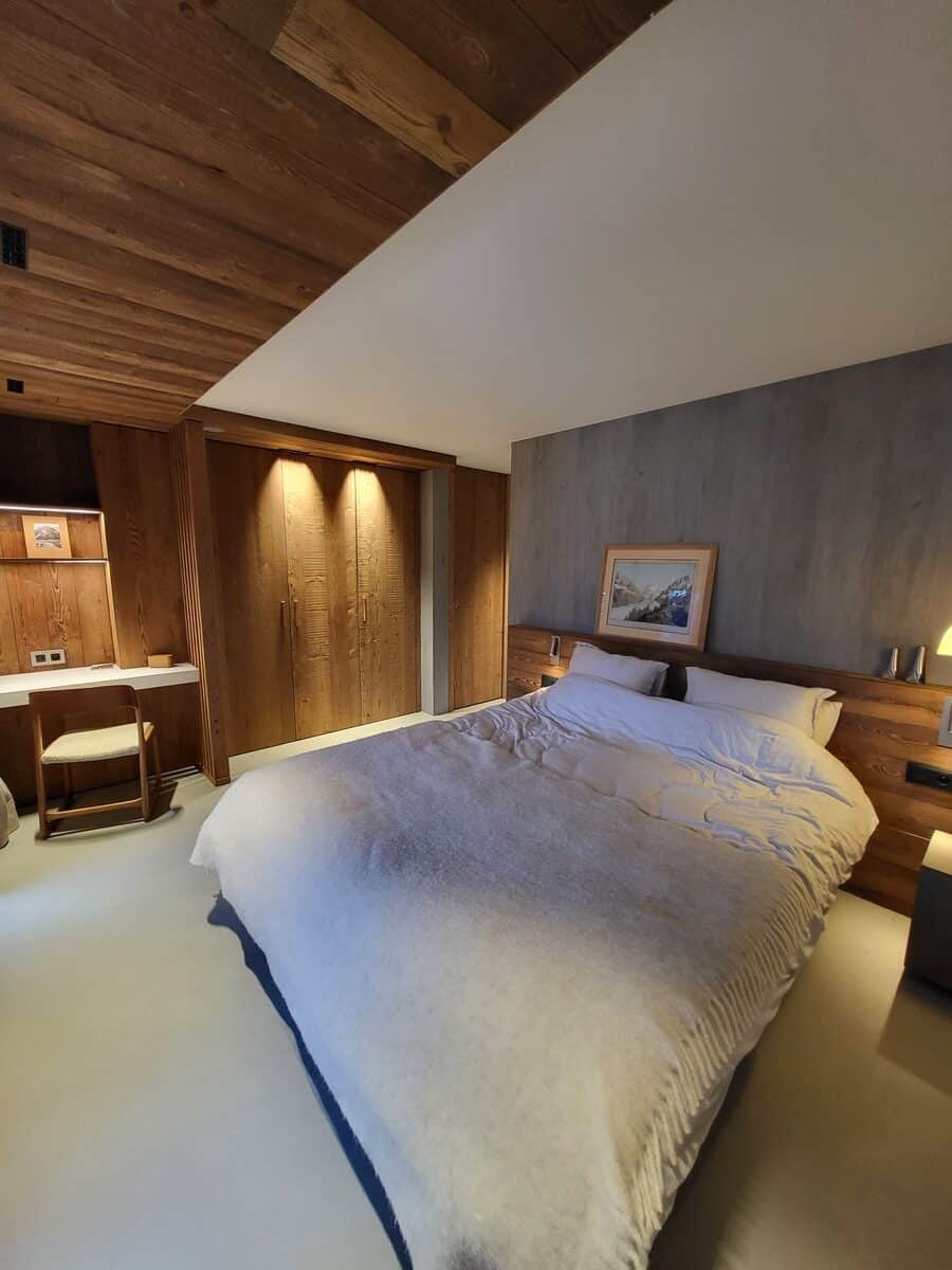 Chambre moderne et confortable. Grand lit double blanc, plafond en bois, murs gris et mobilier boisé. Coin bureau élégant.