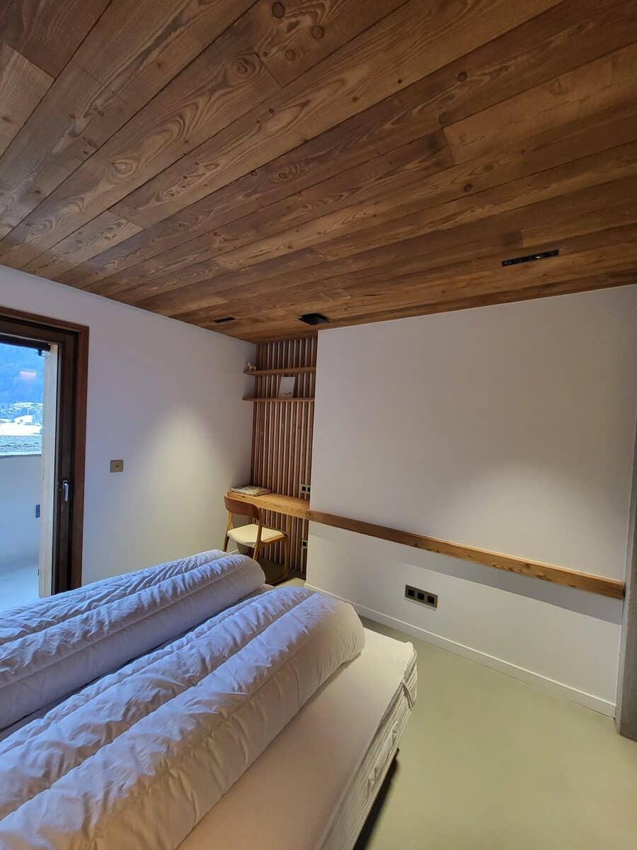 Chambre moderne au plafond bois texturé, lit double blanc et coin bureau intégré. Ambiance chaleureuse avec vue montagne discrète.