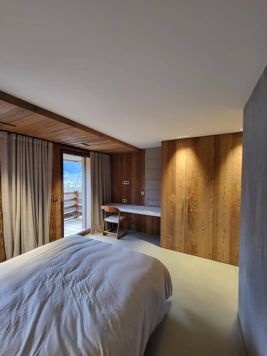 Chambre moderne avec lit, murs en bois clair, bureau intégré et balcon offrant une vue sur les montagnes enneigées.