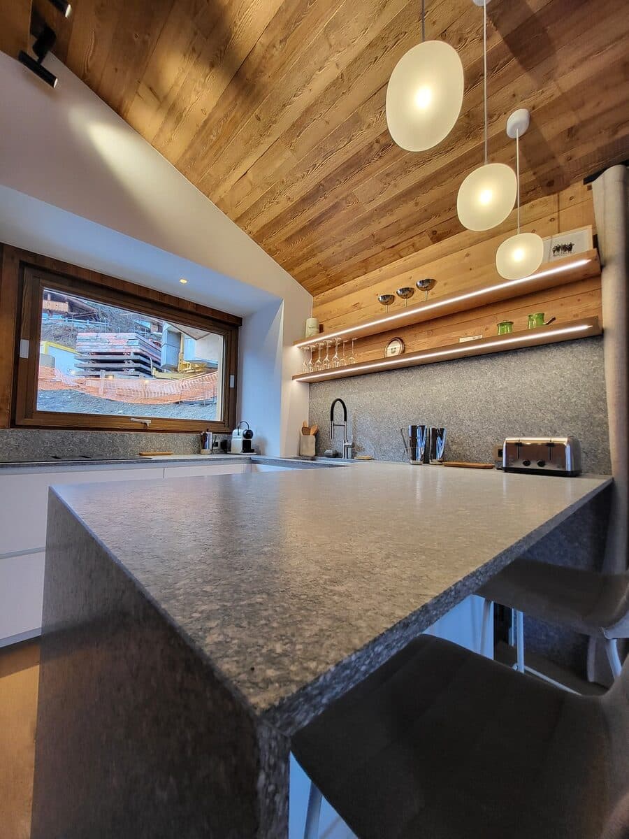 Cuisine moderne avec plafond en bois incliné, îlot central en granit gris, et luminaires suspendus. Fenêtre donnant sur un chantier.