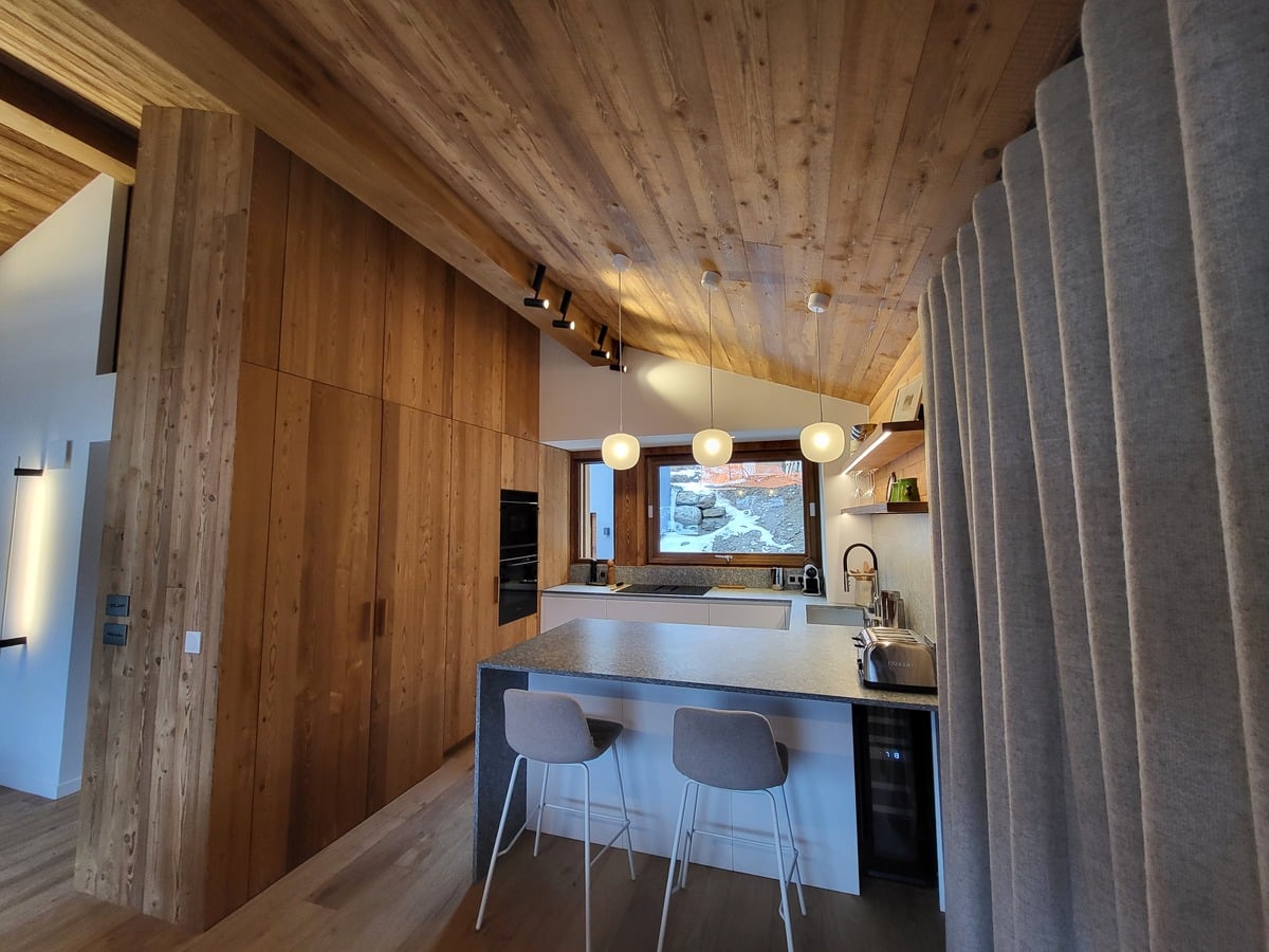 Cuisine moderne avec îlot central, deux tabourets, et mur en bois clair. Plafond en lattes de bois, luminaires suspendus et fenêtre sur paysage enneigé.