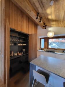 Cuisine moderne avec cellier intégré en bois clair, comptoir et îlot en granit gris, et plafond incliné en bois. Spots et suspension lumineuse.