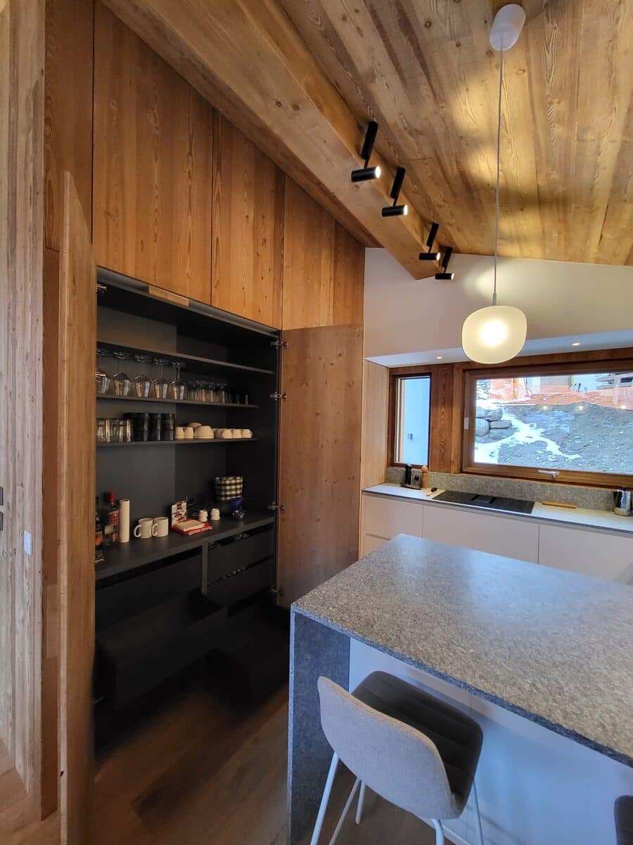 Cuisine moderne avec cellier intégré en bois clair, comptoir et îlot en granit gris, et plafond incliné en bois. Spots et suspension lumineuse.