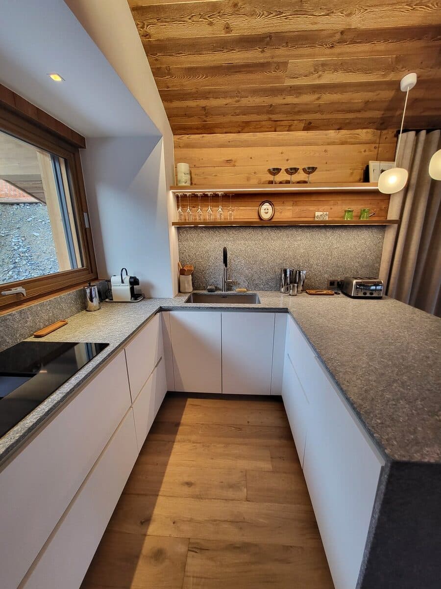 Cuisine moderne en U, meubles blancs sans poignée, plan de travail gris moucheté. Plafond en bois rustique, étagères lumineuses et fenêtre.