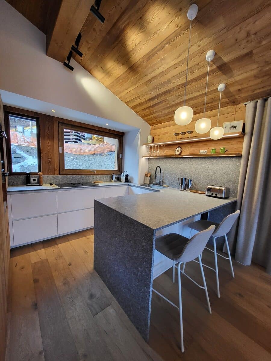 Cuisine moderne style chalet avec plafond en bois, îlot central en granit, tabourets de bar, armoires blanches et grande fenêtre.