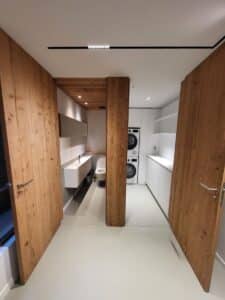 Couloir design avec portes en bois ouvertes. À gauche, salle de bain minimaliste (vasque, WC). À droite, buanderie (lave-linge, sèche-linge, rangements blancs).
