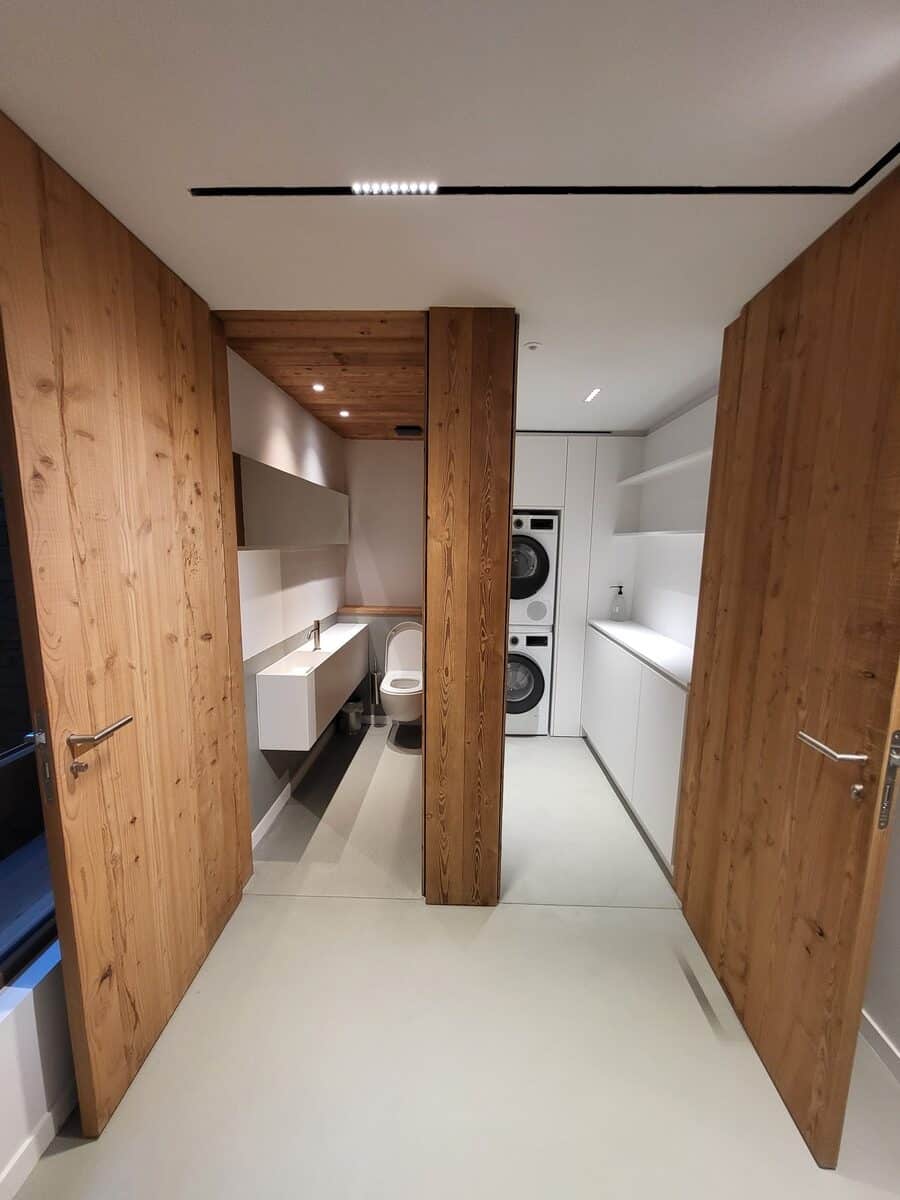 Couloir design avec portes en bois ouvertes. À gauche, salle de bain minimaliste (vasque, WC). À droite, buanderie (lave-linge, sèche-linge, rangements blancs).