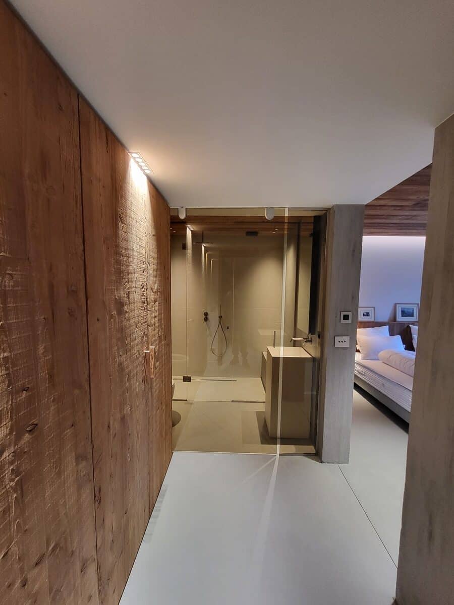 Design moderne: mur en bois, salle de bain vitrée avec douche et lavabo, menant à une chambre lumineuse avec lit. Éclairage discret.