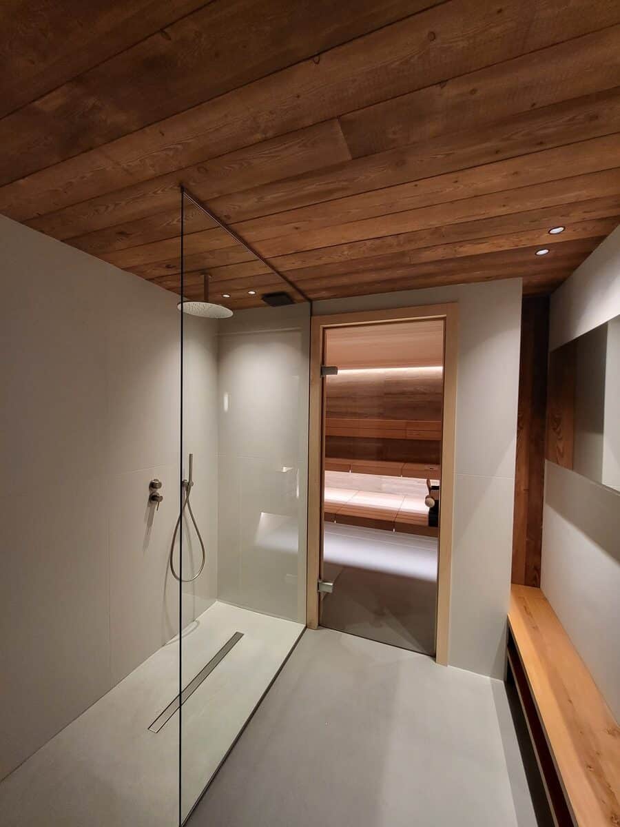 Douche italienne moderne avec paroi vitrée, menant à un sauna en bois. Plafond boisé clair, sol gris. Ambiance spa relaxation.