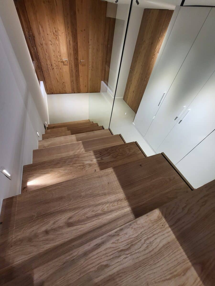 Escalier moderne en bois clair descendant, éclairé par des spots muraux. Garde-corps en verre, placards blancs intégrés et plafond en bois.