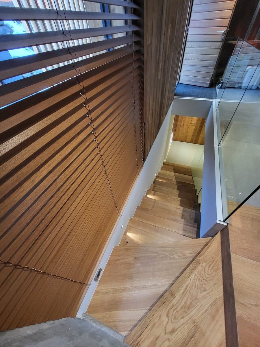 Escalier en bois clair moderne avec balustrade en verre et lumières intégrées, sous des stores vénitiens foncés filtrant la lumière dans un intérieur design.