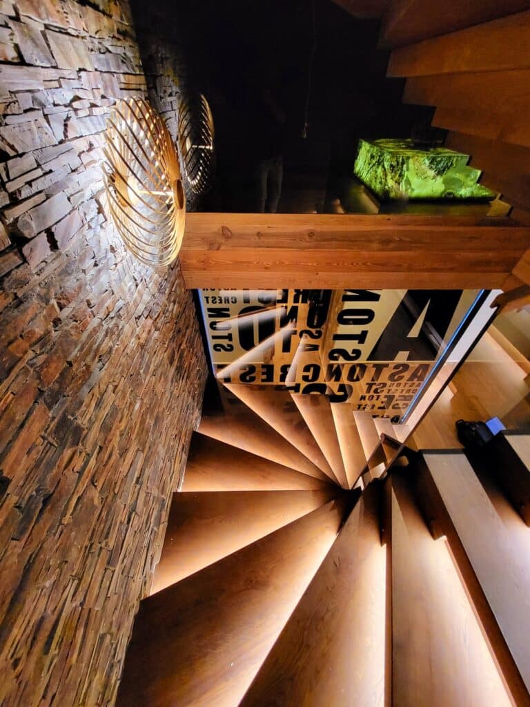 Escalier colimaçon en bois clair avec éclairage intégré, longeant un mur en pierres. Deux luminaires design spiralés décorent le mur. Reflets de texte : ASTON, GUEST, LOW, CHEST.