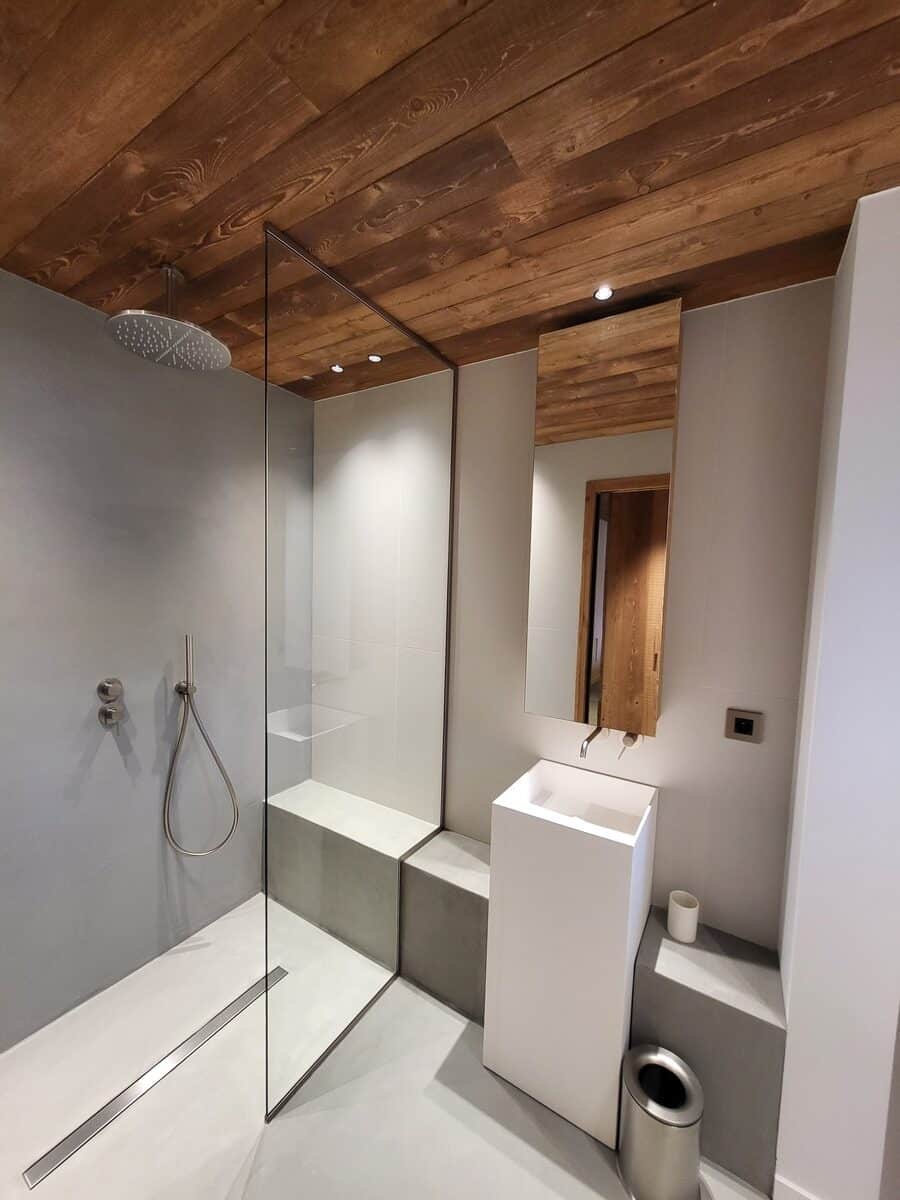 Salle de bain moderne minimaliste : plafond bois, douche à l'italienne verre et béton, lavabo colonne blanc design, miroir et accessoires argentés.
