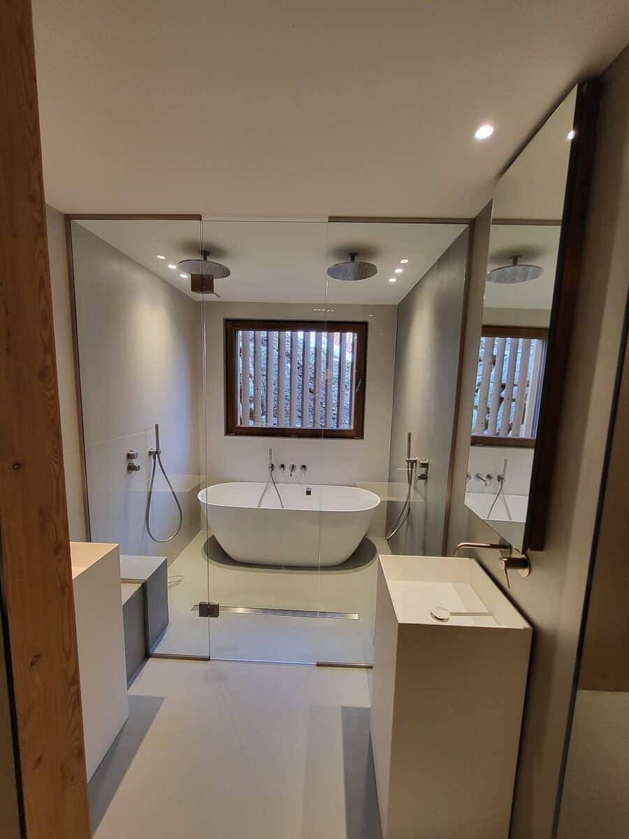 Salle de bain moderne avec baignoire îlot, deux douches pluie intégrées, lavabos colonne blancs et paroi vitrée.