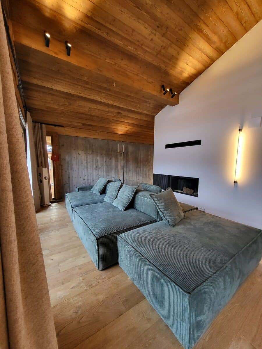 Grand canapé modulable vert-de-gris côtelé dans un salon moderne. Plafond en bois, mur en béton et cheminée linéaire sur mur blanc.