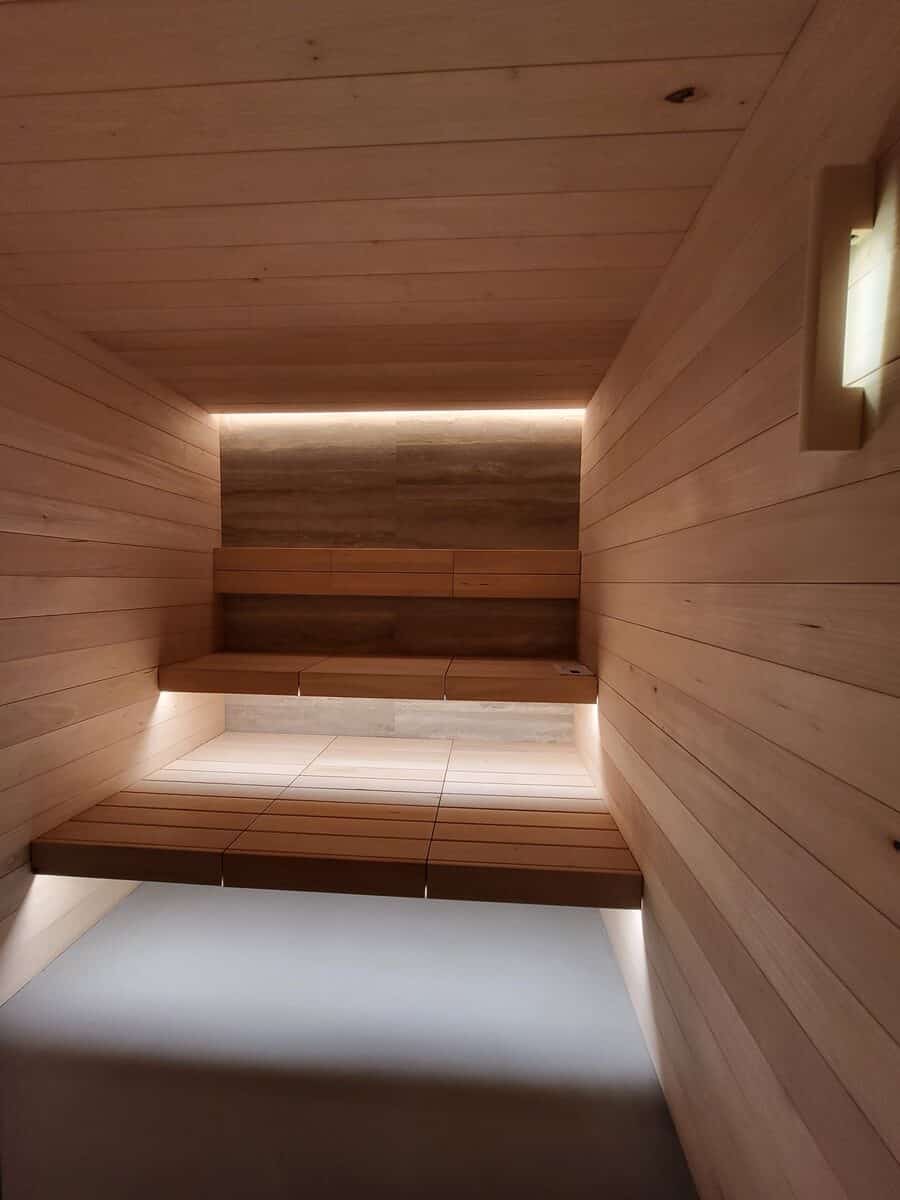 Sauna moderne en bois clair avec bancs étagés et éclairage LED indirect. Mur du fond texturé et design épuré.
