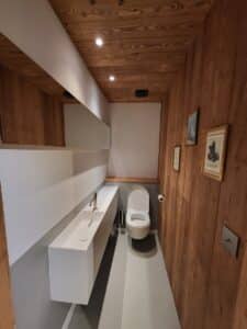 WC design avec murs et plafond en bois. Long lavabo blanc suspendu, miroir, toilettes modernes et sol gris.