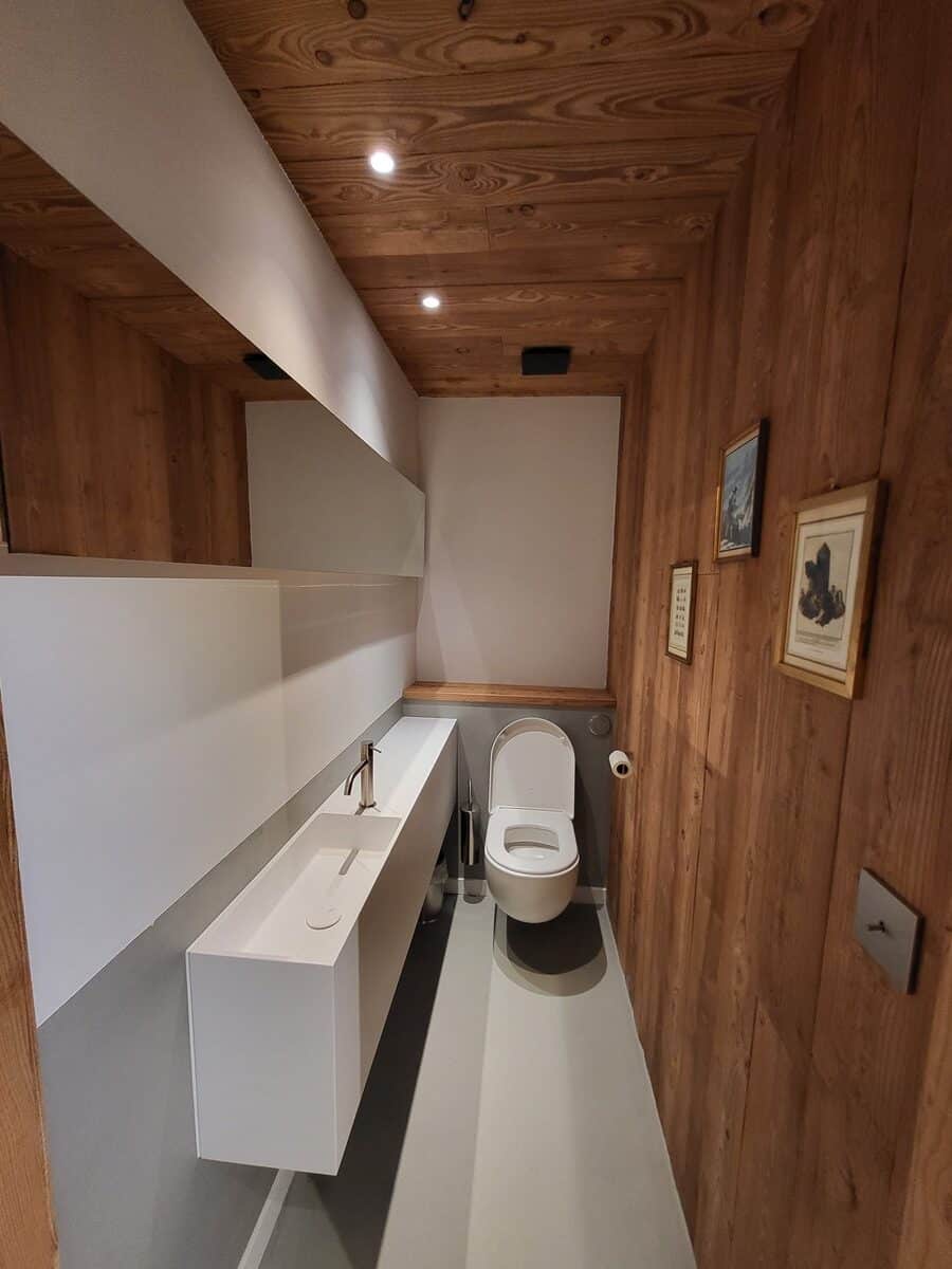 WC design avec murs et plafond en bois. Long lavabo blanc suspendu, miroir, toilettes modernes et sol gris.