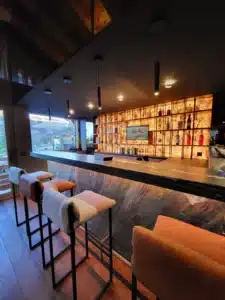 Bar moderne avec comptoir en pierre sombre, tabourets colorés, et étagères rétroéclairées en onyx remplies de bouteilles d'alcool.