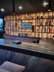 Bar moderne avec étagère rétroéclairée en onyx présentant des bouteilles d'alcool, comptoir en pierre sombre et caisson noir marqué 'COGNAC'.