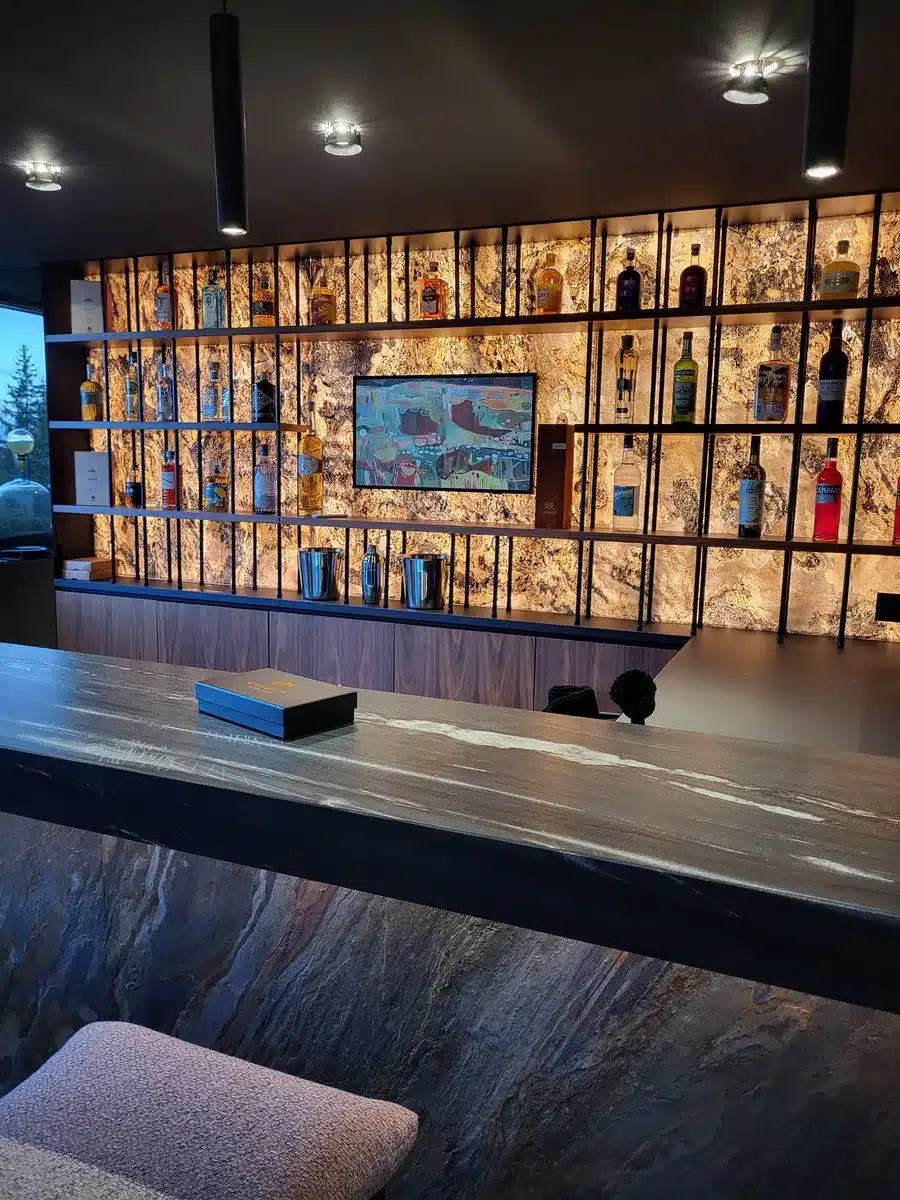Bar moderne avec étagère rétroéclairée en onyx présentant des bouteilles d'alcool, comptoir en pierre sombre et caisson noir marqué 'COGNAC'.