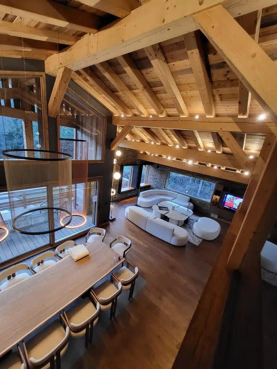 Vue plongeante d'un grand chalet moderne avec poutres apparentes, salle à manger boisée et salon blanc contemporain, lumière chaude.