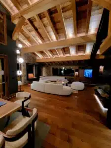 Salon alpin luxueux avec plafond en poutres bois apparentes, canapé blanc incurvé, murs en pierre et parquet brun riche.