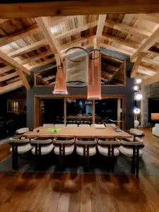 Salle à manger de chalet luxueux avec plafond cathédrale en bois rustique, grande table rectangulaire et chaises modernes blanches éclairées par des suspensions design.
