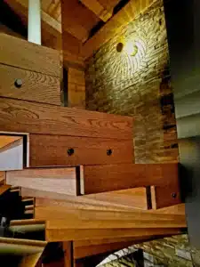 Escalier flottant en bois de chêne richement veiné, éclairé par une applique murale sculpturale sur un mur de pierre rustique.