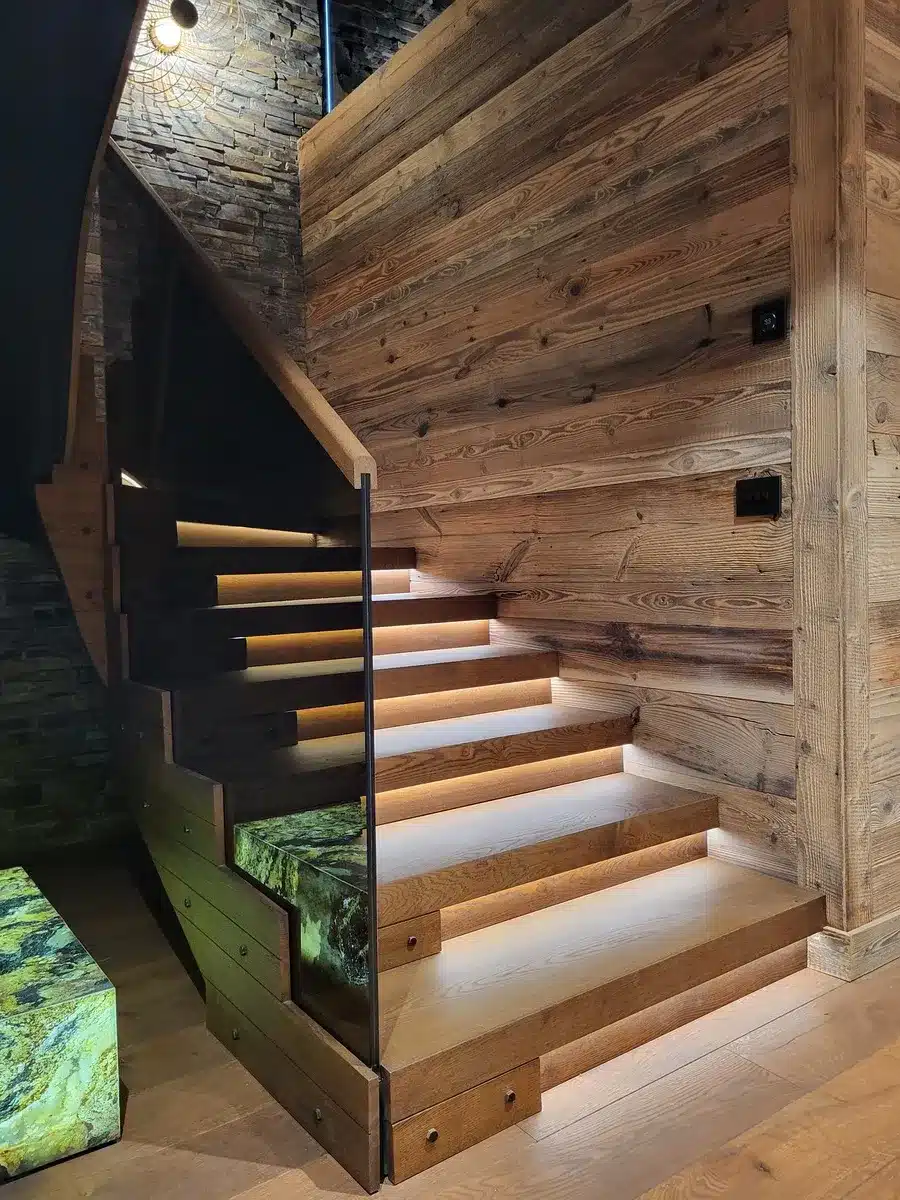 Escalier moderne en bois avec éclairage LED sous les marches, garde-corps en verre et mur de bardage rustique.