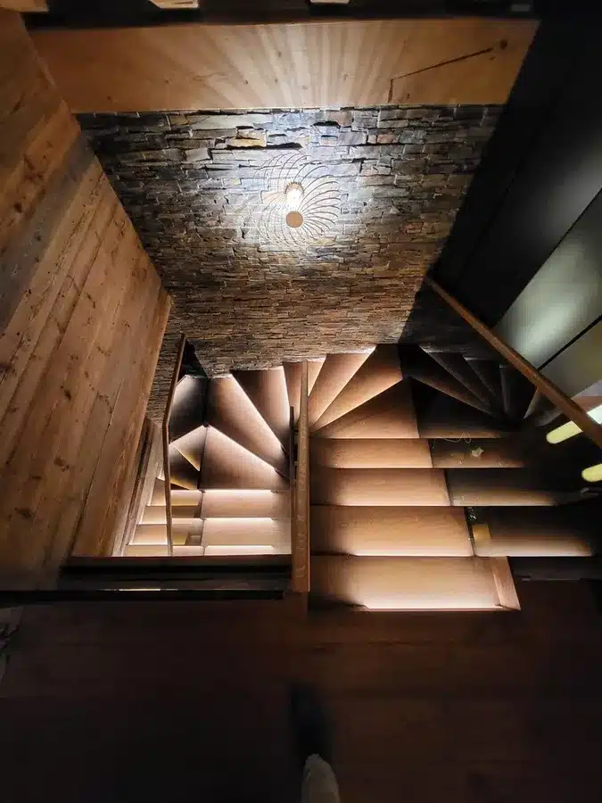 Escalier en bois moderne et chaleureux éclairé par des rubans LED sous les marches, menant à un palier en pierre sèche et un luminaire suspendu.
