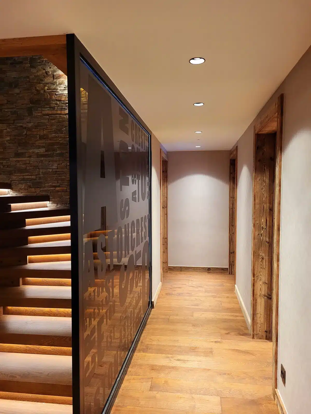 Hall moderne avec sol en bois clair, escalier éclairé, cloison vitrée texturée avec 'ASTONCREST', et portes encadrées de bois brut.